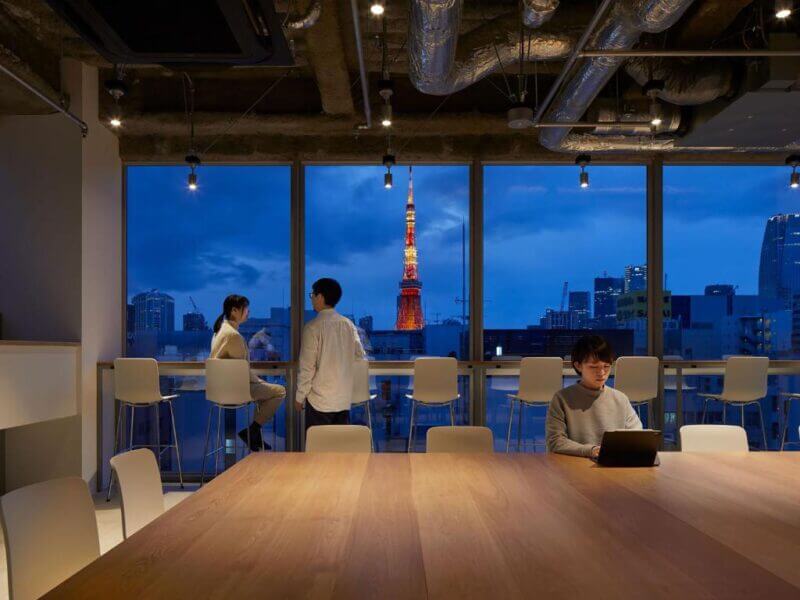 Three people are in a modern office with large windows overlooking a cityscape at dusk, featuring a brightly lit tower and the sleek design reminiscent of Nine Hours Tokyo. One person is using a laptop at the table.