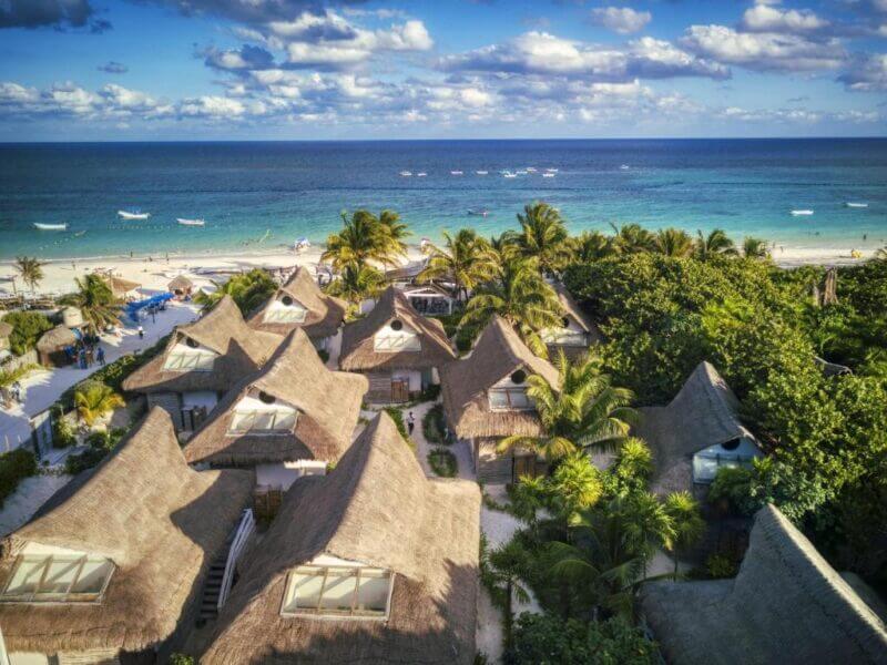 Aerial view of thatched-roof villas at Kai Tulum, surrounded by lush greenery near a sandy beach with blue ocean and boats under a partly cloudy sky.