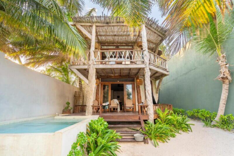 Two-story wooden beach villa with thatched roof, balcony, and shaded patio, surrounded by palm trees and tropical plants. This Casa Ganesh Tulum accommodation features a small pool in front on sandy ground.