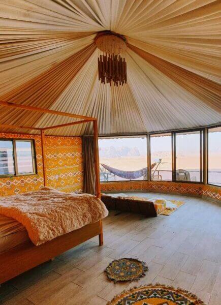 A spacious yurt-style bedroom with a canopy bed, floor rugs, patterned walls, large windows, and stunning views of the Wadi Rum desert landscape—an inviting retreat in a luxurious Desert Camp.