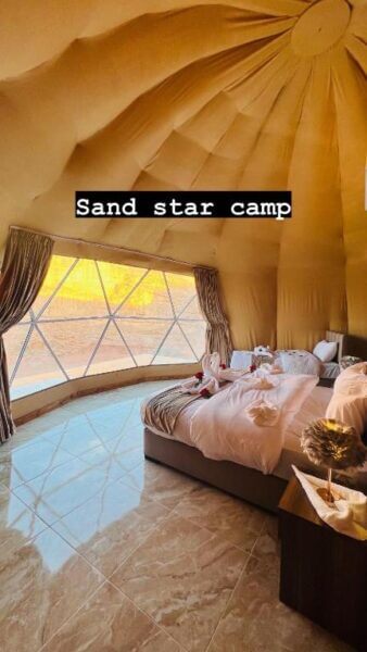 A spacious dome-shaped room with a large bed, big window showing a stunning Wadi Rum desert view, and Sand Star Camp text overlayed at the top.