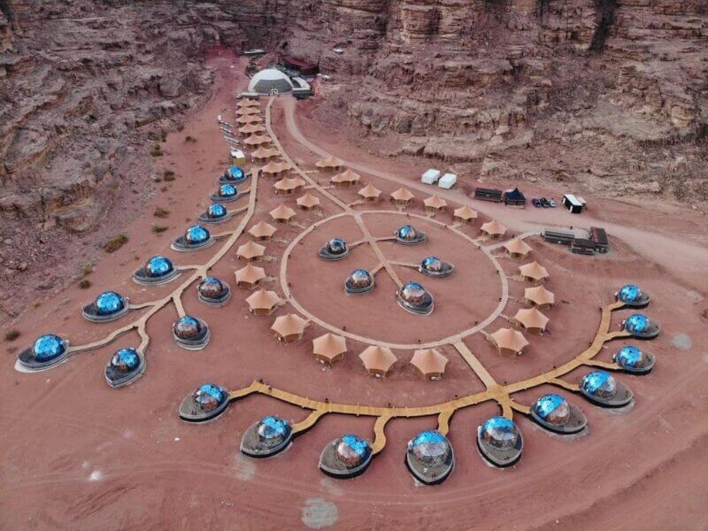 Aerial view of Aicha Luxury Camp, a luxury camp in Wadi Rum, with dome-shaped structures and tan tents arranged in circular patterns amid rocky desert terrain.