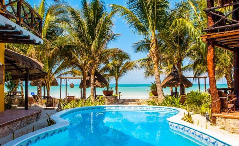A swimming pool surrounded by palm trees at Holbox Dream, a beachfront hotel, with a stunning view of the beach and ocean in the background on a sunny day.