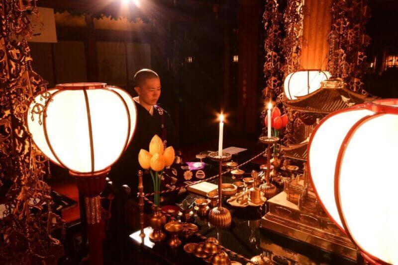 A monk stands at an ornate altar with lit candles and traditional objects, illuminated by large lanterns in a dimly lit room at Yochi-in, a historic Shukubo temple lodging in Koyasan.