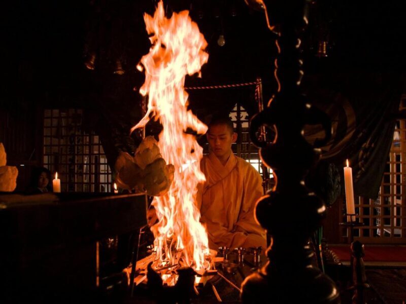 A Buddhist monk in a yellow robe sits behind a tall ceremonial fire inside a dimly lit room with candles and ritual objects at Ekoin Temple, experiencing the sacred traditions of Koyasan’s Syukubo lodging.