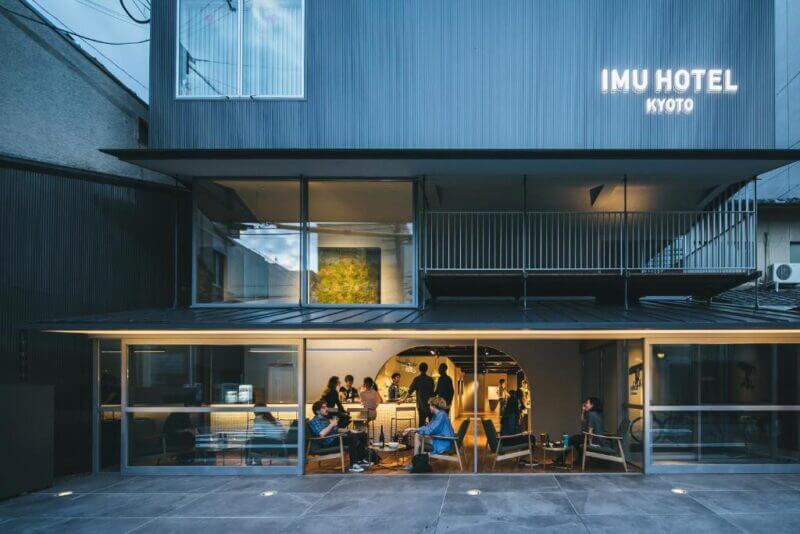 Exterior view of Imu Hotel Kyoto at dusk, with people sitting and socializing on the ground floor patio under warm lighting. Imu Hotel Kyoto’s inviting ambiance creates a perfect spot to unwind in the city.