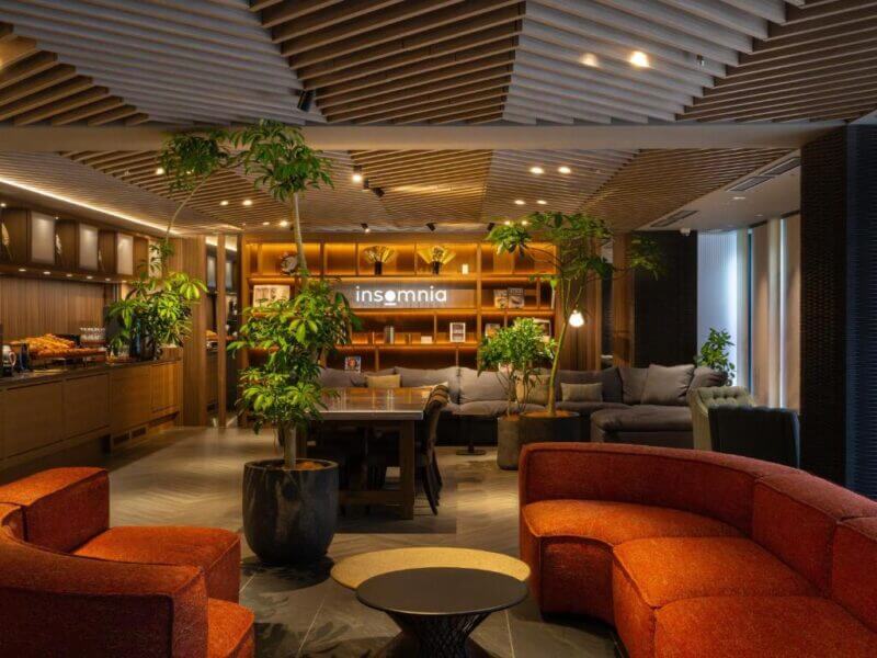 Modern lounge with red curved sofas, potted plants, wooden ceiling panels, and a backlit insomnia sign on bookshelves. Inspired by Kyoto’s serene aesthetics, warm lighting creates a cozy atmosphere perfect for unwinding.