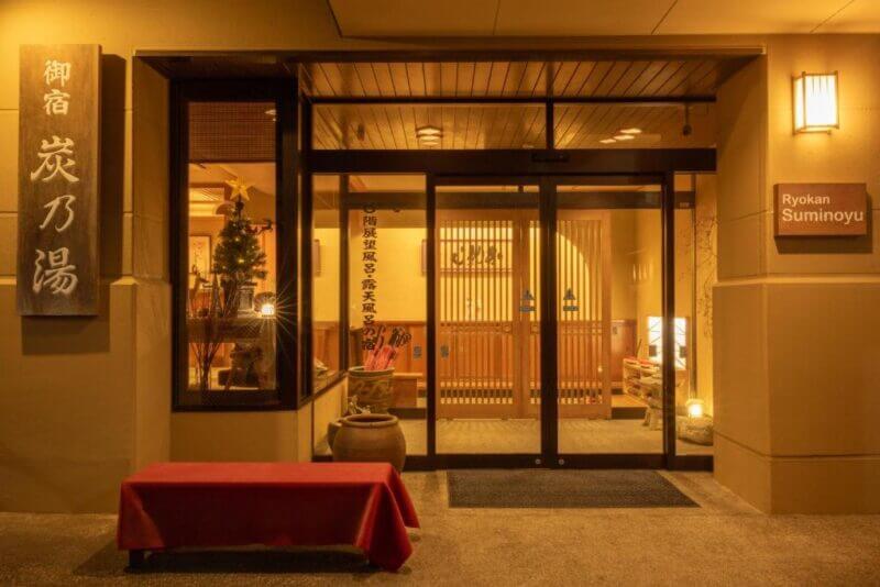 Entrance of Suminoyu Ryokan in Shibu Onsen at night, featuring sliding glass doors, warm lighting, a red bench, and traditional Japanese decor.