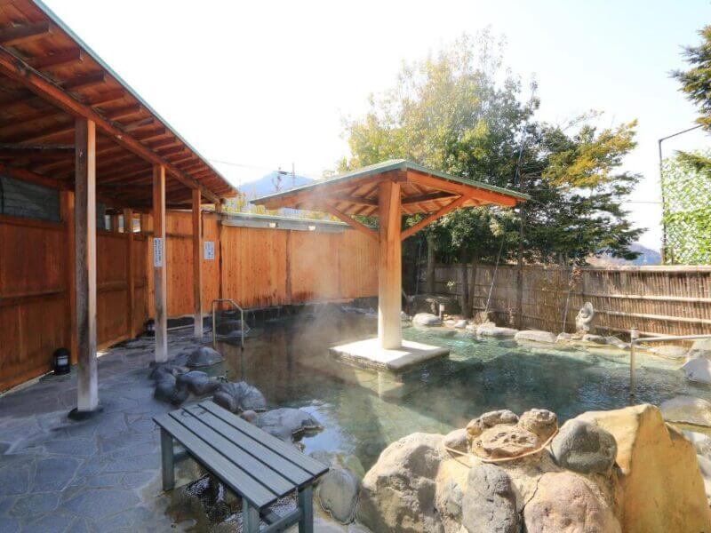 An outdoor Japanese onsen at Minshuku Miyama Shibu with steaming hot spring water, surrounded by rocks, wooden walls, and a wooden canopy structure. A bench sits invitingly on the stone-paved edge.