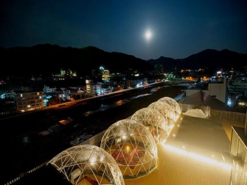 Nighttime view of a city with illuminated transparent dome structures on a rooftop terrace at Aburaya Tousen Yudanaka Ryokan, overlooking the river and mountains under a full moon in the background.