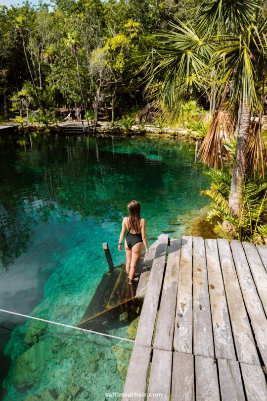 A woman in a swimsuit walks down wooden steps into the clear, blue-green waters of Cenote Corazon Tulum, a hidden gem surrounded by trees and tropical plantsâ€”one of the top things to do in Yucatan, Mexico.