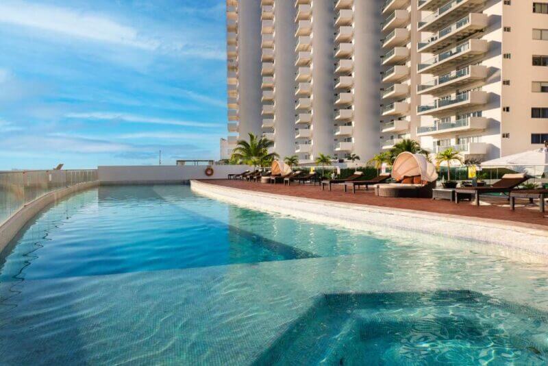Curved outdoor swimming pool with lounge chairs and umbrellas beside the tall, modern Cancun Suites hotel building under a blue sky near the vibrant Malecon.
