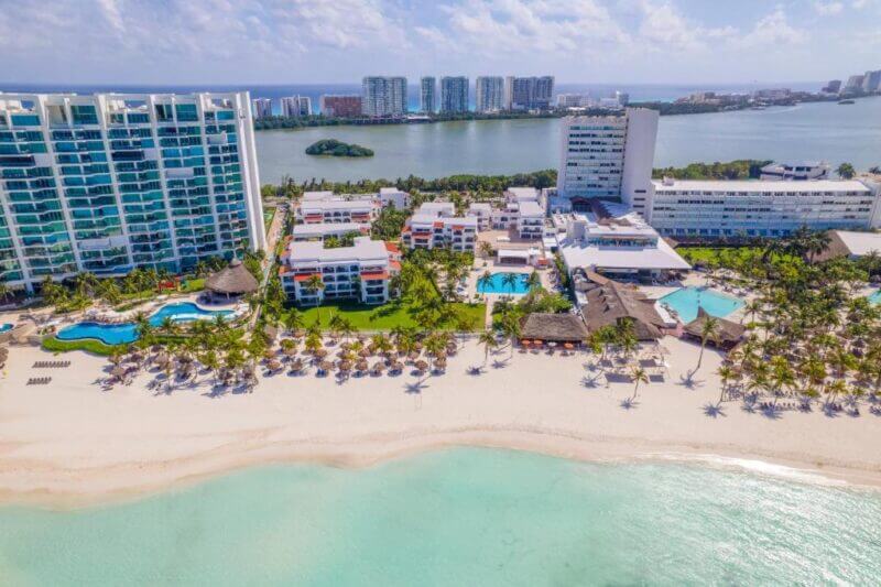 Aerial view of Beachscape Kin Ha Cancun, featuring pools, palm trees, and lounge chairs along white sand and turquoise water; stylish Villas & Suites overlook the lagoon with city buildings in the background.