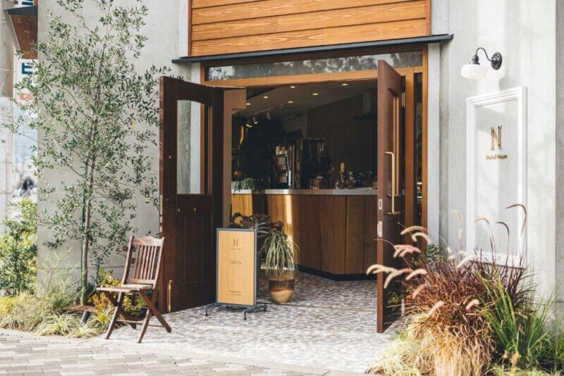 A cafe near Hotel Noum OSAKA with open wooden doors, a small sign, a chair outside, and plants around the entrance. The interior features a counter and warm lighting.