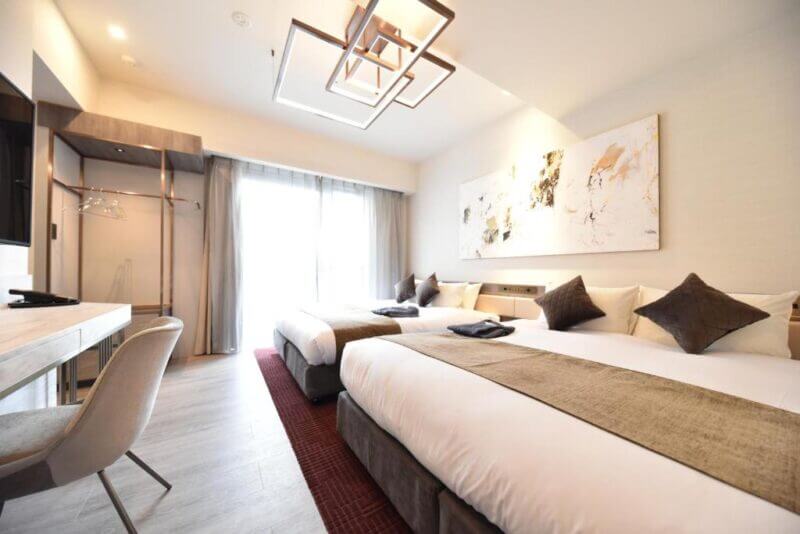 Modern Hotel Cordia Osaka room with two large beds, a desk and chair, floor-to-ceiling window with curtains, and abstract wall art above the beds.