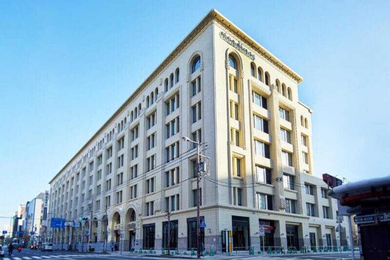 A large, beige, multi-story building with arched windows on the ground floor, Citadines Namba Osaka is a serviced apartment located on a city street corner under a clear blue sky.