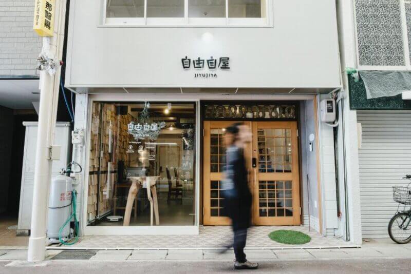 A person walks past the glass-front entrance of Jiyuya, a modern store in Nachikatsuura called 自由自屋, with shelves and wooden furniture visible inside.