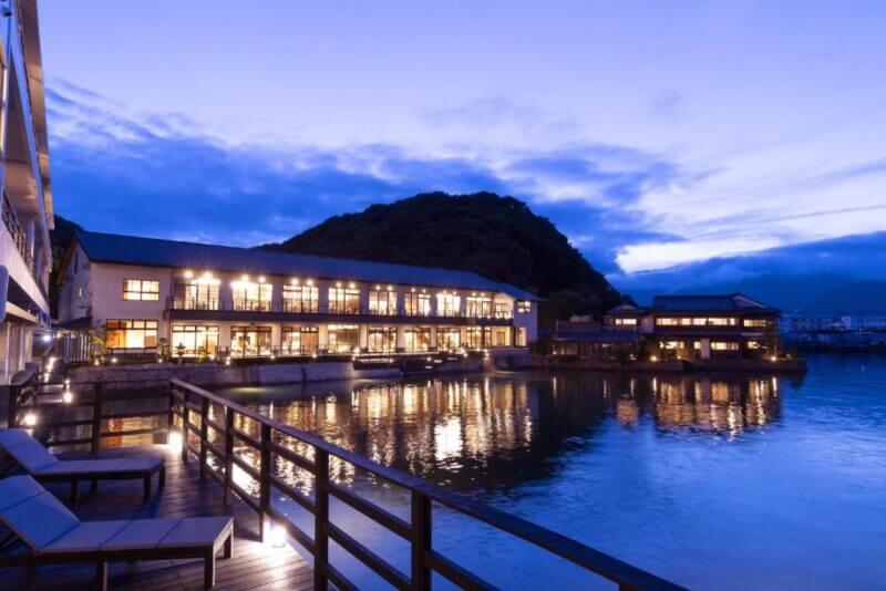 A waterfront resort with outdoor lounge chairs and illuminated windows, nestled by a calm body of water at dusk in Nachikatsuura, with a tree-covered hill providing a scenic backdrop.