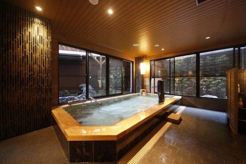 A modern indoor Japanese-style hot spring bath at Onyado Nono Nara, featuring large windows, a wooden ceiling, stone tile flooring, and warm ambient lighting for a serene stay at this renowned Nara hotel.