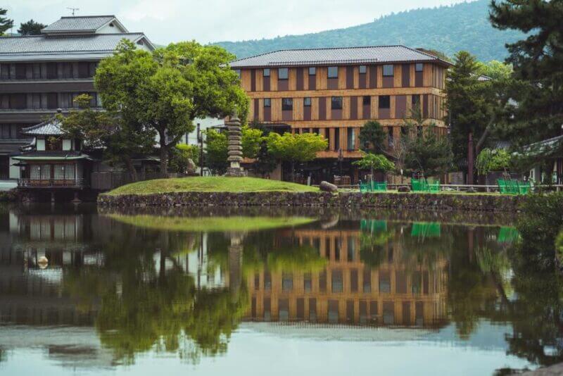 A traditional Japanese building and stone pagoda at SETRE Naramachi hotel are reflected in a calm pond, surrounded by trees and the mountains of Nara in the background.