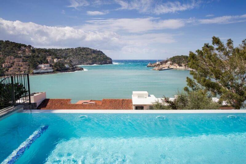 Infinity pool at Hotel Esplendido overlooking Soller Bay with turquoise water, coastal buildings, and hills under a partly cloudy sky.