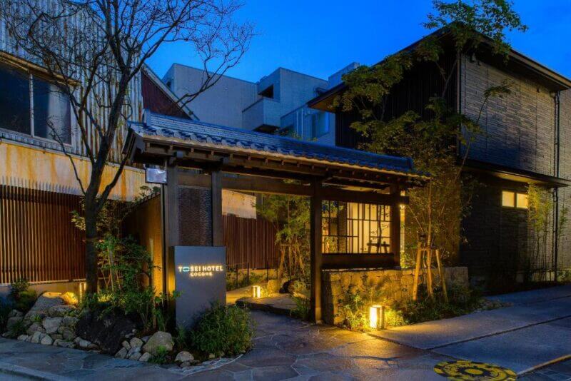 A traditional Japanese-style hotel entrance with a wooden gate, garden lights, and modern buildings in the background at dusk welcomes you to Hotel Cocone Kamakura.