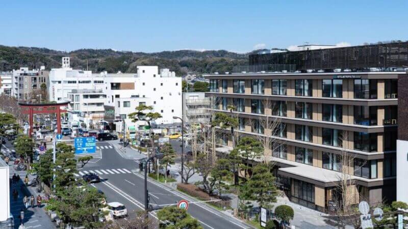 A city street scene in Kamakura shows modern buildings, cars, pedestrians, and a large red torii gate in the background under a clear sky, with the Kamakura hotel and Hotel Metropolitan nearby.