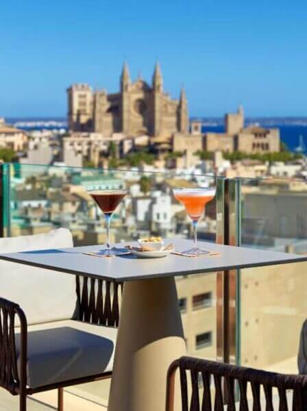 A rooftop table at Hotel Saratoga with two cocktails and snacks overlooks Palma’s cityscape, featuring a large historic cathedral under a clear blue sky.