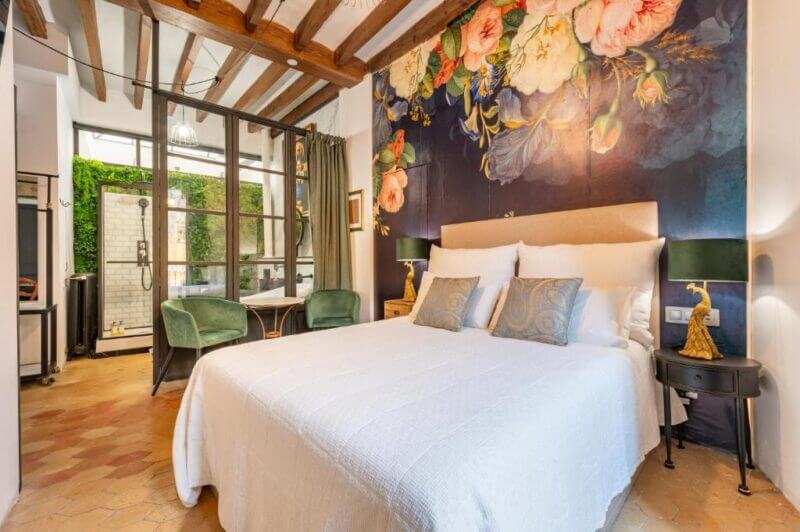 A bedroom in Palma with a large bed, floral mural accent wall, wooden ceiling beams, green chairs, and a glass door leading to an outdoor area—perfect for relaxing with your dog.