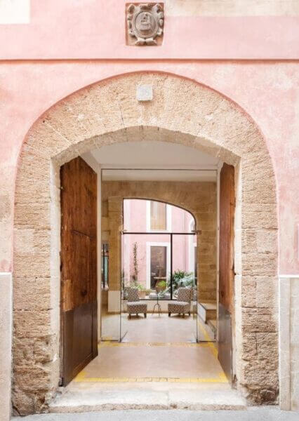 Stone archway with wooden doors opens to an indoor courtyard at Palau Sa Font in Palma, featuring potted plants, chairs, and pink walls with large windows in the background.
