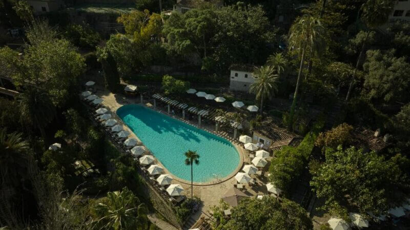 Aerial view of Es Moli Deia’s large outdoor swimming pool, surrounded by lounge chairs, umbrellas, palm trees, and dense greenery.