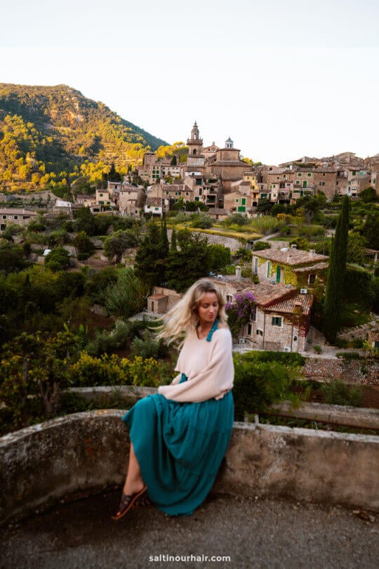 A woman in a teal skirt and light sweater sits on a stone ledge in Valldemossa, Mallorca, with a hillside village and lush greenery of the Balearic Islands in the background at sunrise.
