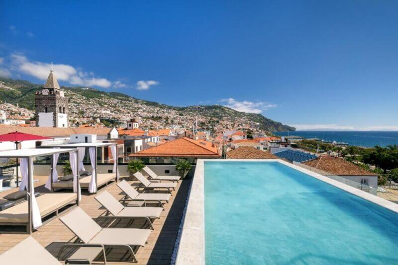 Rooftop infinity pool at Barceló Funchal with lounge chairs overlooking Madeira’s cityscape, mountains in the background, and the ocean on the right under a clear blue sky.