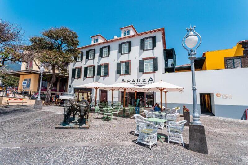 Outdoor cafe seating with white umbrellas and green chairs sits in front of a three-story building at Viola Market Studios in Funchal. A street lamp and bronze statues enhance the charm of the cobblestone plaza.