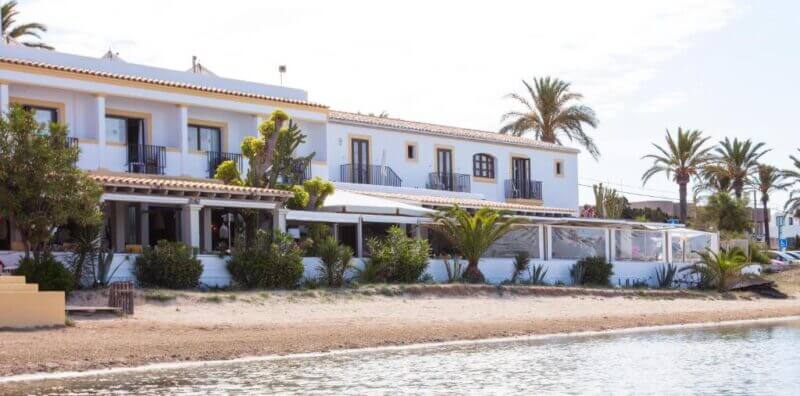 Hostal la Savina is a white two-story accommodation with balconies and palm trees, located directly on a sandy beach beside calm water in beautiful Formentera.