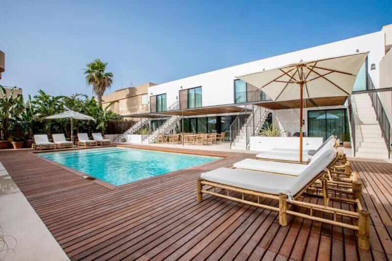 Modern outdoor pool area at Can Micalet Formentera, featuring a wooden deck, white lounge chairs, umbrellas, and a two-story building with glass doors in the background.