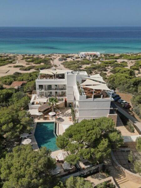 A modern white Teranka Formentera hotel with a pool is surrounded by trees, set near a sandy beach and blue sea under a clear sky.