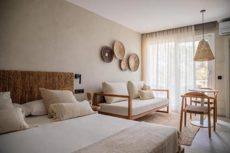 A modern bedroom at Boho Suites Formentera featuring a double bed, a sofa, a small table with two chairs, woven decor on the wall, and large windows with sheer curtains letting in natural light.