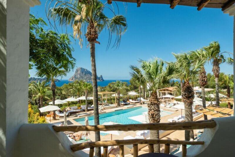 Enjoy the stunning view from your balcony at Petunia Hotel Ibiza, overlooking a sparkling swimming pool, swaying palm trees, sun loungers, and the ocean with a rocky island under a clear blue sky.