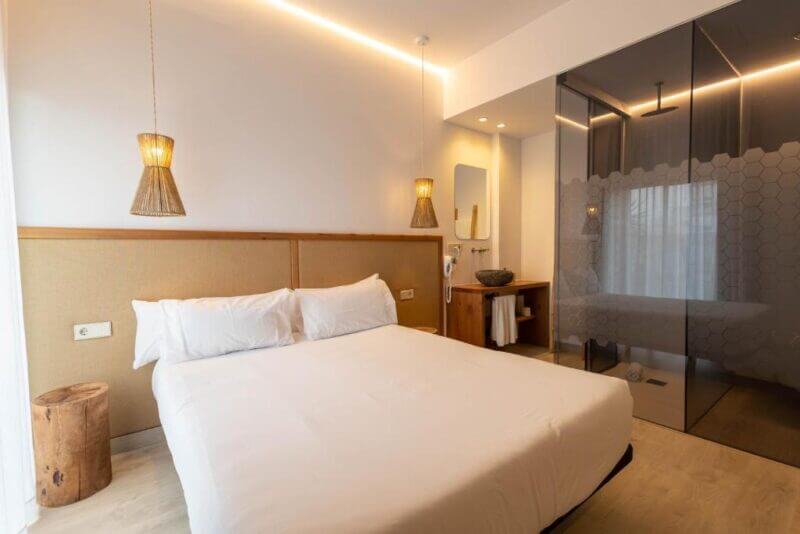 Minimalist Hotel Ibiza room with a double bed, two hanging wicker lamps, a small wooden stool, and an open bathroom area featuring glass walls and a rain shower.