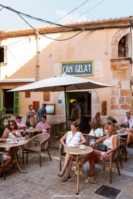 People sit at outdoor tables eating and drinking under umbrellas in front of a shop called Can Gelat, enjoying the lively atmosphere near the Santanyi Market Mallorca on a sunny day.