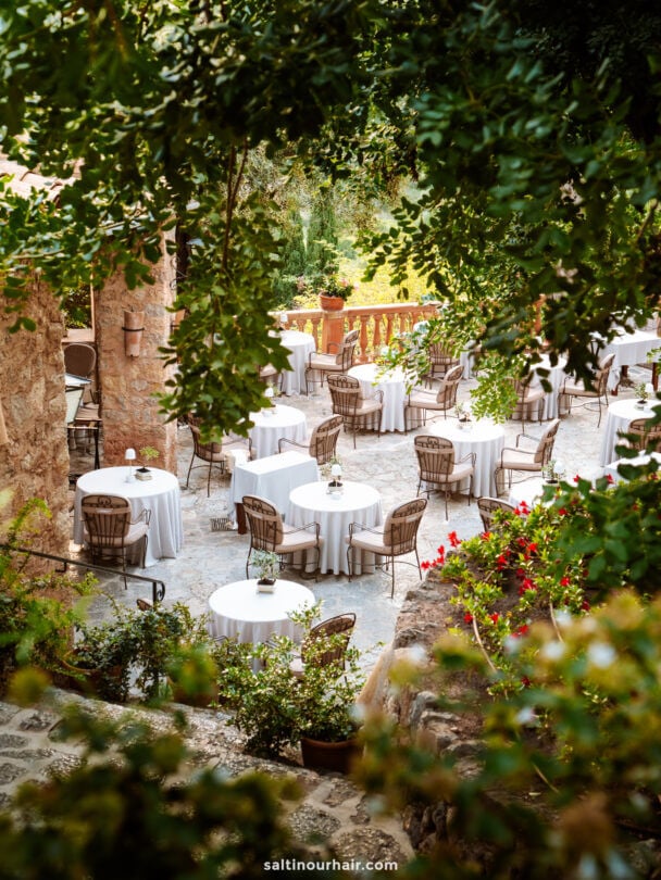 Outdoor restaurant with round tables covered in white tablecloths, surrounded by chairs, set among stone buildings and lush greenery in the charming village of Deia, Mallorca, Balearic Islands.