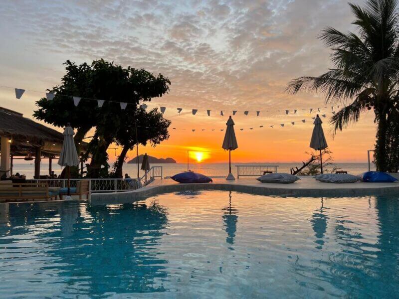 A poolside scene at sunset in Koh Phangan with lounge chairs, umbrellas, and string lights; silhouettes of trees and the sun setting over the ocean in the background.