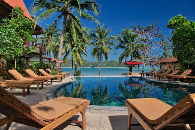 A swimming pool surrounded by wooden lounge chairs and palm trees at Longtail Beach Resort overlooks a calm beach and blue ocean under a clear sky on Koh Phangan.