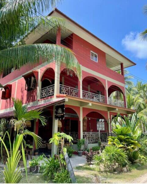A three-story red guesthouse with multiple arches and balconies, surrounded by tropical plants under a blue sky with scattered clouds—perfect for a relaxing stay in Koh Phangan.