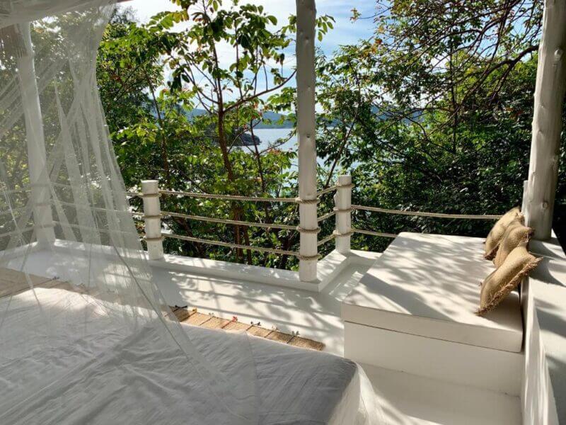 A white outdoor bed with mosquito net at Coconut Beach Bungalows faces a railing and lush green trees, with glimpses of water and blue sky in the background. Two tan pillows rest on a white bench—perfect for relaxing on Koh Phangan.
