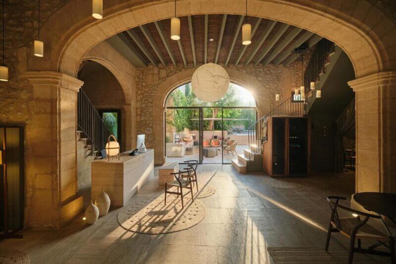 Spacious room with stone arches, wooden ceiling, and modern furniture at Can Aulí in Pollenca, opening to an outdoor patio with seating and greenery visible through large glass doors.