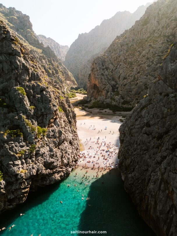Aerial view of Sa Calobra Beach, Mallorca, nestled between steep rocky cliffs in the Balearic Islands, with people swimming and sunbathing by turquoise water.