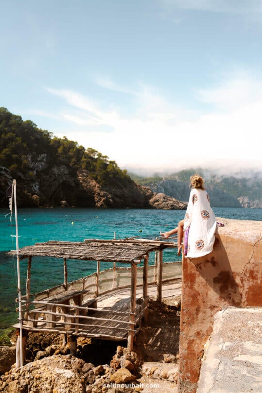 A woman wearing a white cover-up sits on a stone wall overlooking clear turquoise water, a wooden dock, and tree-covered hillsâ€”perfect inspiration for things to do in Ibiza under a partly cloudy sky.