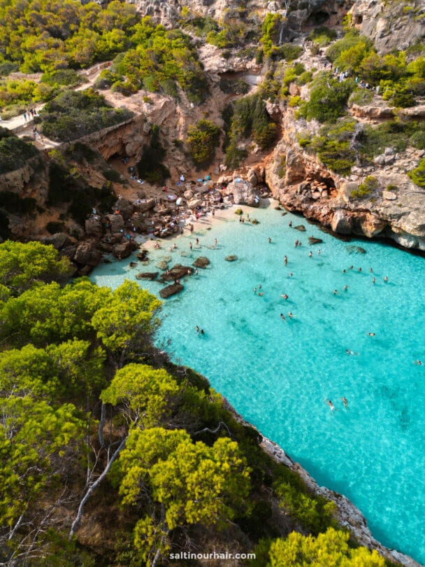 Aerial view of Cala del Moro, Mallorca, Spain: a turquoise cove surrounded by rocky cliffs and dense green trees, with people swimming and sunbathing on the beach.